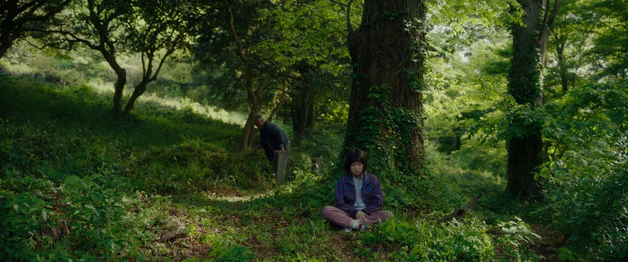 Okja (2017) movie still: wide — A quiet clearing within a dense woodland area featuring old stone grave markers. The girl sits against a large tree…