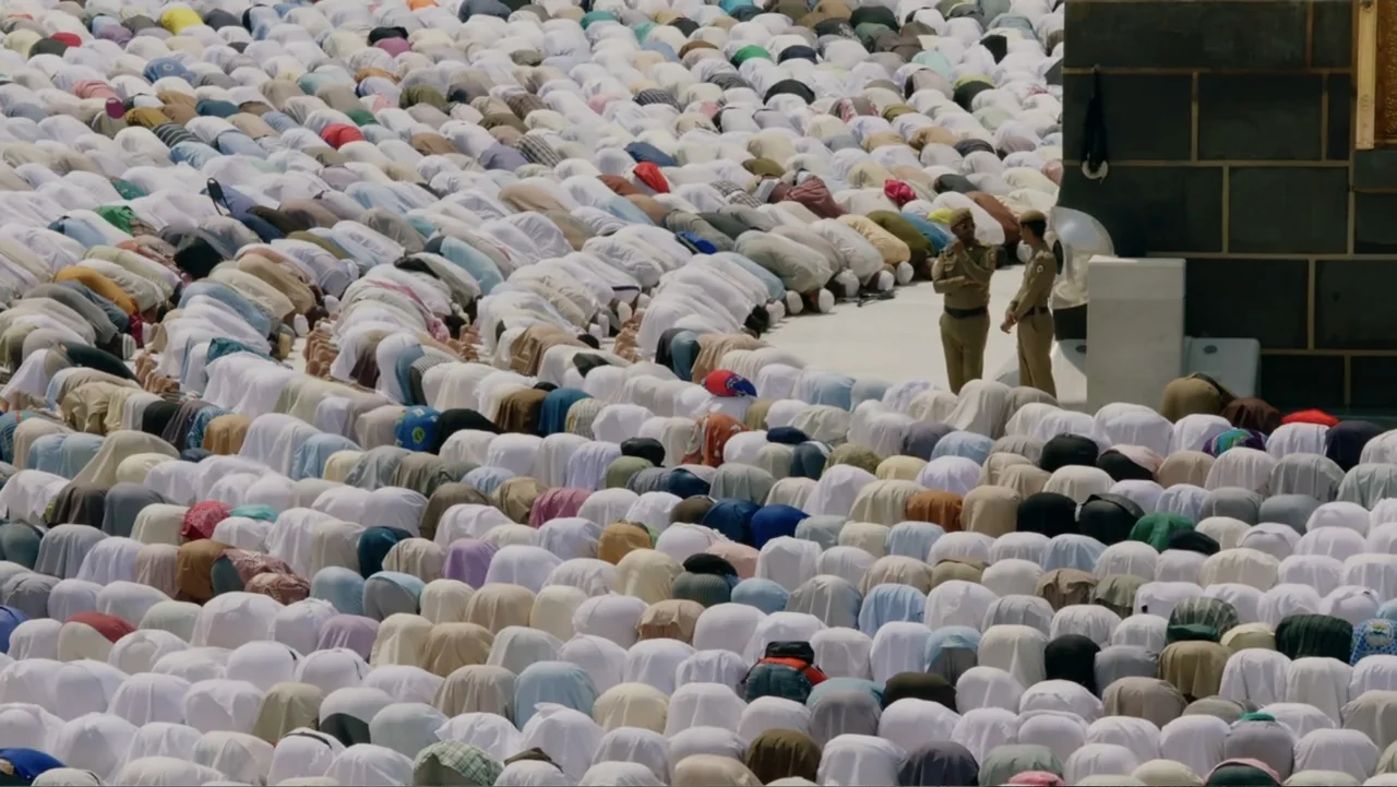 One Day in the Haram (2017) movie still: wide — A vast marble plaza is filled with a dense crowd kneeling in unison. Two security guards stand near…