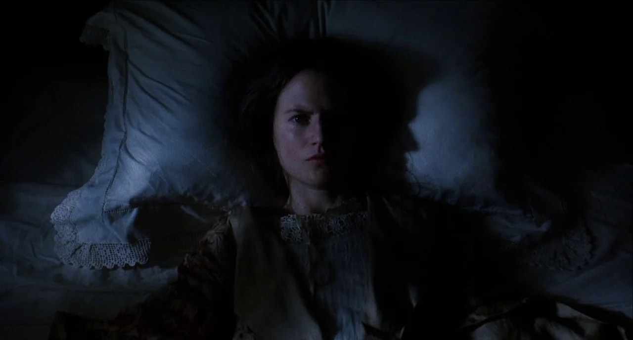 The Hours (2002) movie still: closeup — A woman lies flat in bed against white linens with lace trim at night. She is positioned frontally with her…
