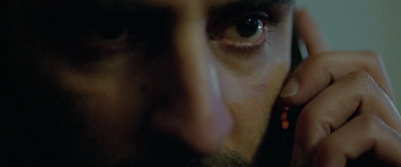 Catch Me Daddy (2014) movie still: closeup — A man's face is partially obscured by shadows in an dimly lit environment. He holds a phone against his…