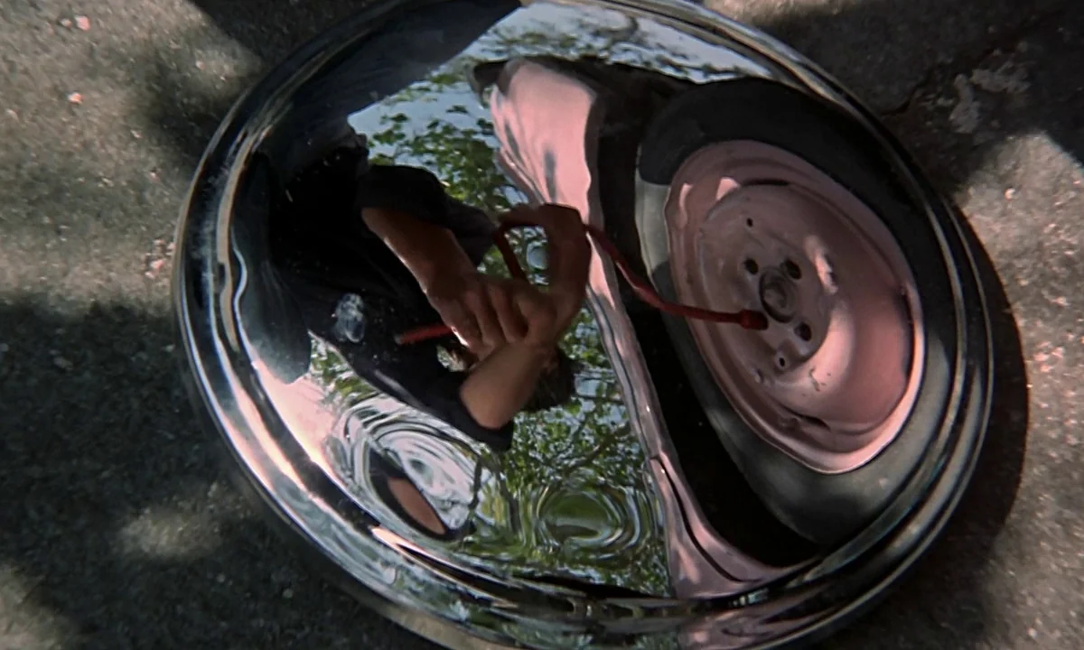 Help! (1965) movie still: closeup — A shiny circular hubcap sits on a piece of grey asphalt pavement. The hubcap reflects a person pumping air into…