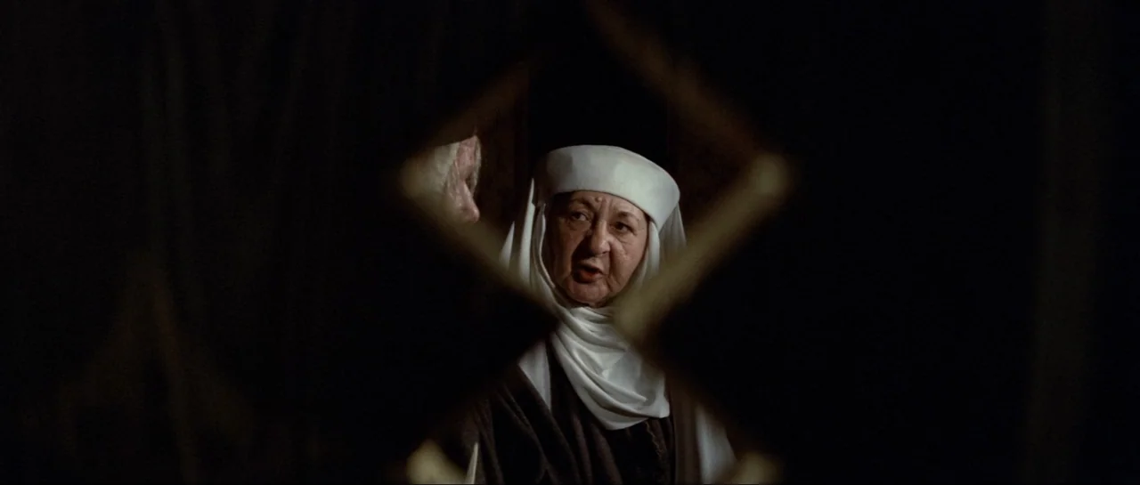 Macbeth (1971) movie still: closeup — A dark stone room is obscured by a diamond-shaped cutout in a wooden screen. An older woman wearing a white…