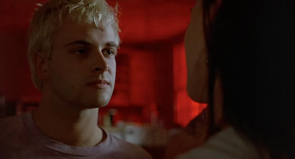Trainspotting (1996) movie still: closeup — A dimly lit interior space is bathed in intense red light from an unseen source. A young man with…