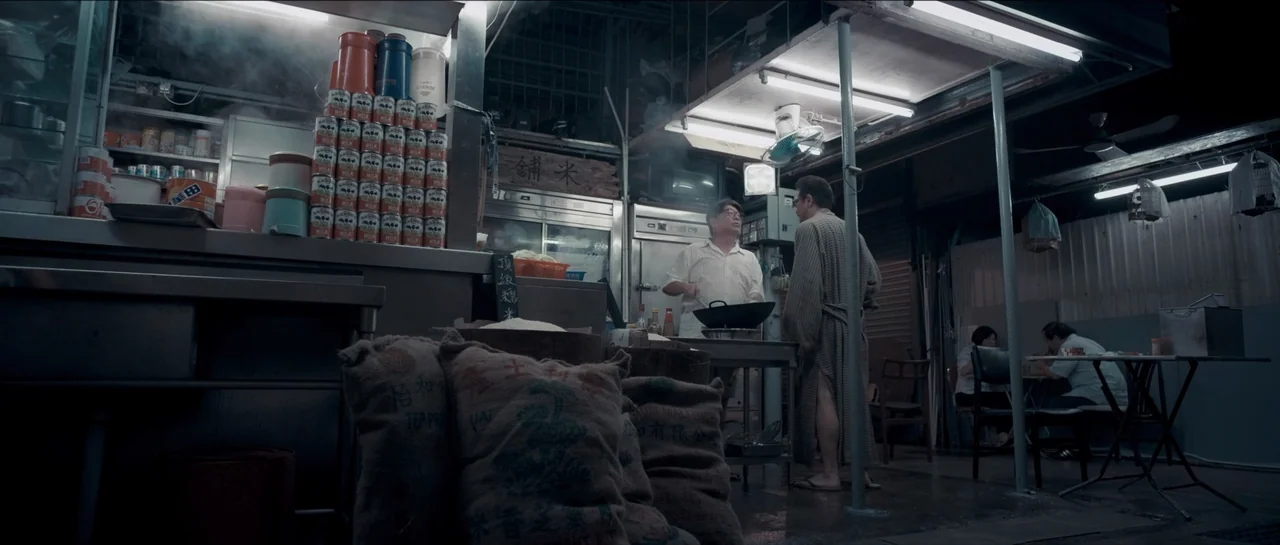 Rigor Mortis (2013) movie still: wide — A nighttime open-air food stall features stacks of canned goods and heavy burlap sacks. A cook in a white…