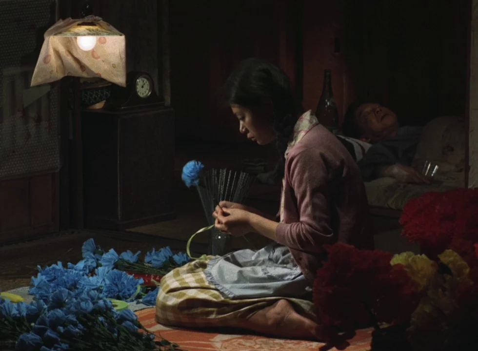 Dodes’ka-den (1970) movie still: medium — A dimly lit room contains a woman crafting artificial flowers on the floor. An older man rests on a mat in…