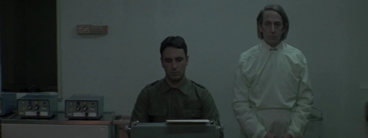 Post Mortem (2010) movie still: medium — A utilitarian room features a typewriter and medical electronics against a blank wall. Two men stand…