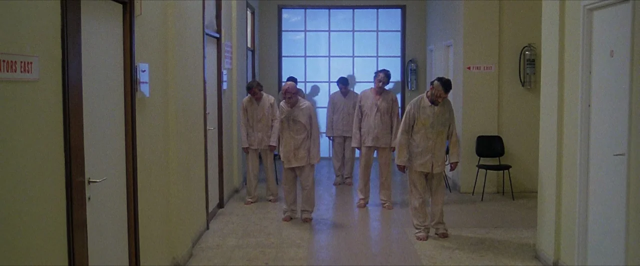 The Beyond (1981) movie still: fullbody — A long beige institutional hallway with white doors and a distant frosted glass entrance. Five men in…
