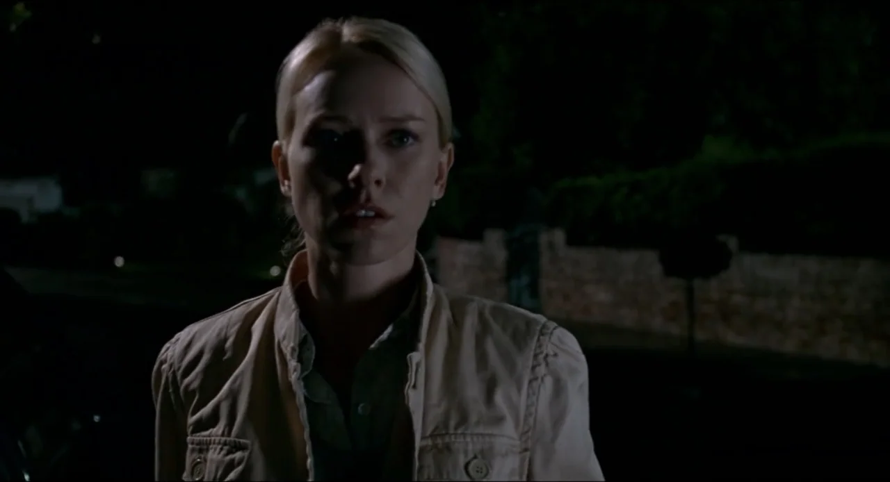 The Ring Two (2005) movie still: medium — An exterior street at night with dark landscaping visible in the background. A woman in a beige jacket…