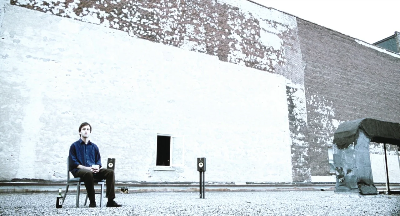 Last Night (1998) movie still: wide — A flat rooftop covered in gravel against a large, weathered, white-painted brick wall. A man sits alone on a…