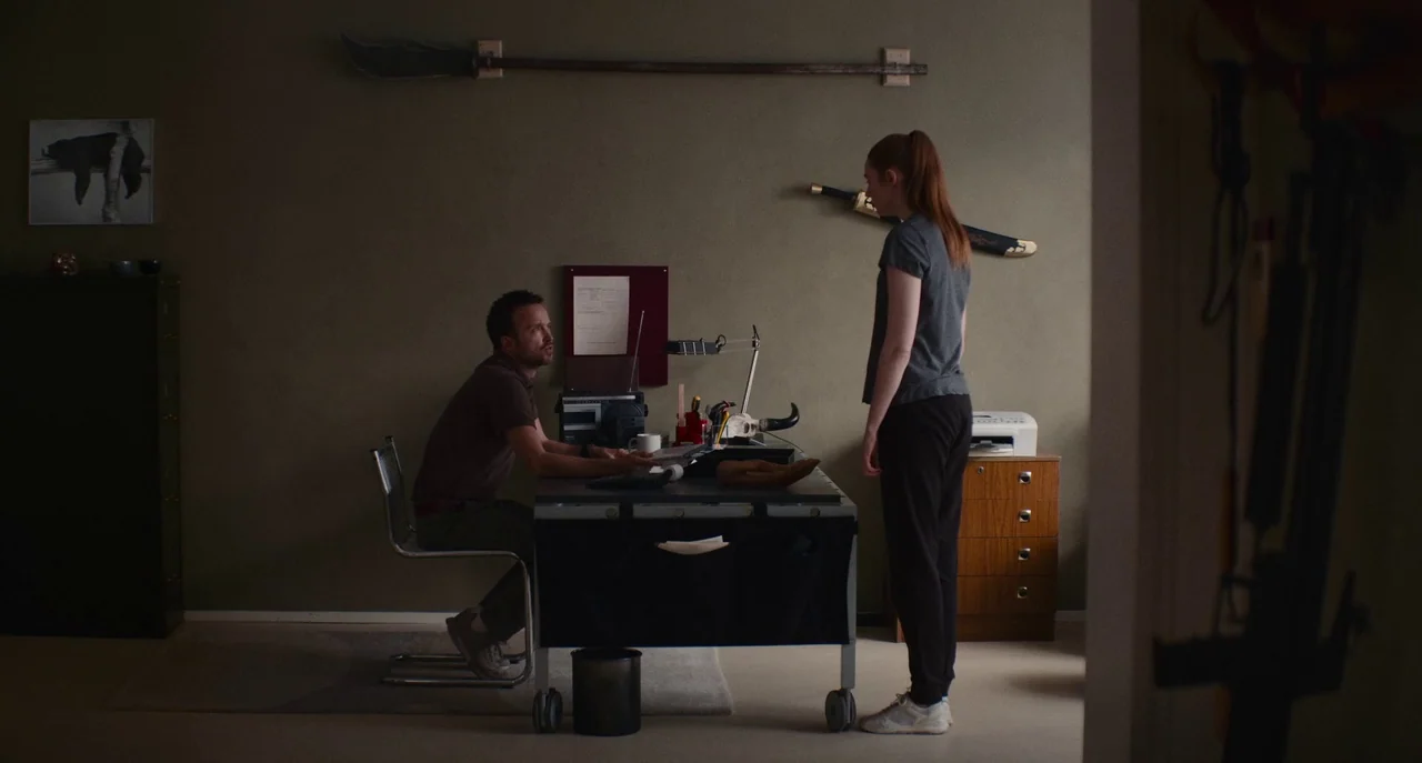 Dual (2022) movie still: wide — A spare, muted office contains a metal desk, a filing cabinet, and wall-mounted weapons. A man sits at the desk…