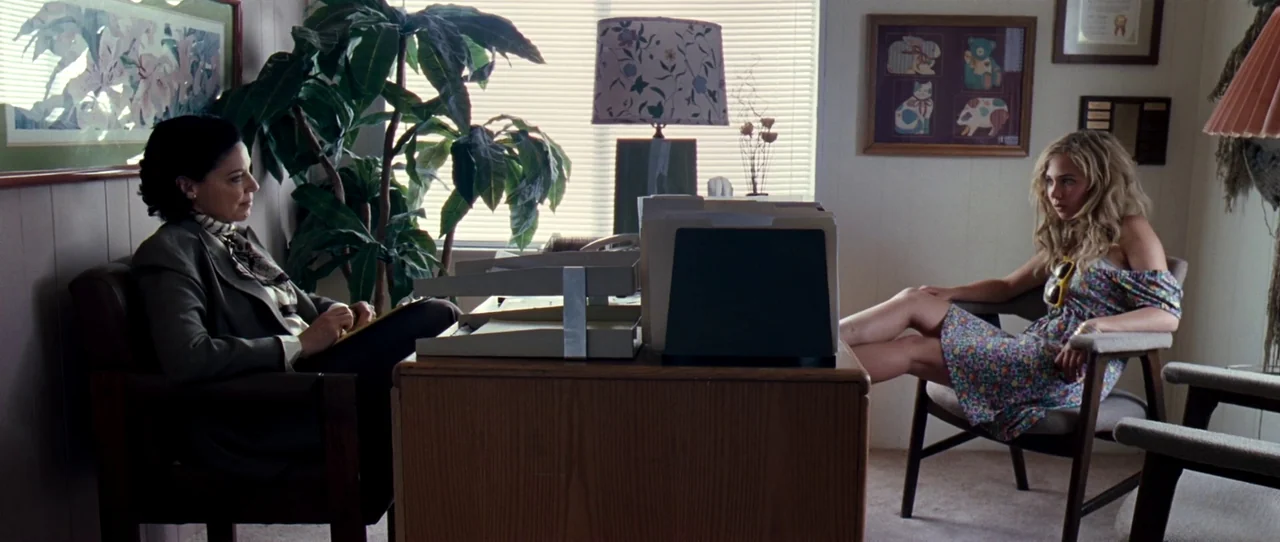 Little Birds (2011) movie still: wide — An office features paneled walls and a wooden desk with a file organizer. An older woman faces a younger…