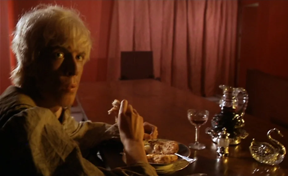 The Immortal Story (1968) movie still: medium — A dimly lit room features walls draped in heavy red fabric curtains. A young man sits at a dark…