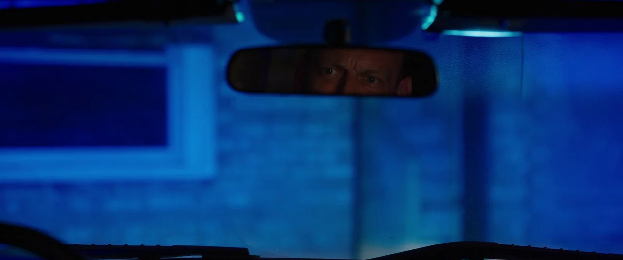 Candyman (2021) movie still: closeup — The interior of a vehicle is bathed in deep blue neon light at night. A man's eyes are visible in the center…