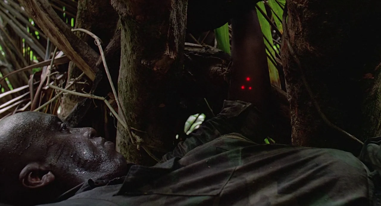 Predator (1987) movie still: closeup — A man lies prone among thick tree trunks and dense green jungle foliage. Three red laser dots are visible on…