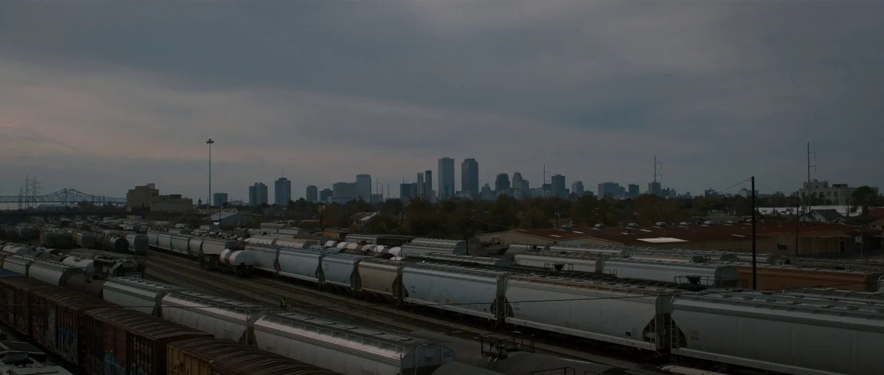 The East (2013) movie still: establishing — A vast railway yard filled with numerous white freight cars stretches toward a distant city. Under a…