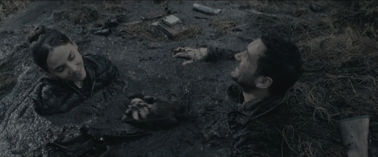 Quicksand (2023) movie still: medium — A man and woman are trapped in a thick, dark mud pit outdoors. They are partially submerged and look toward…