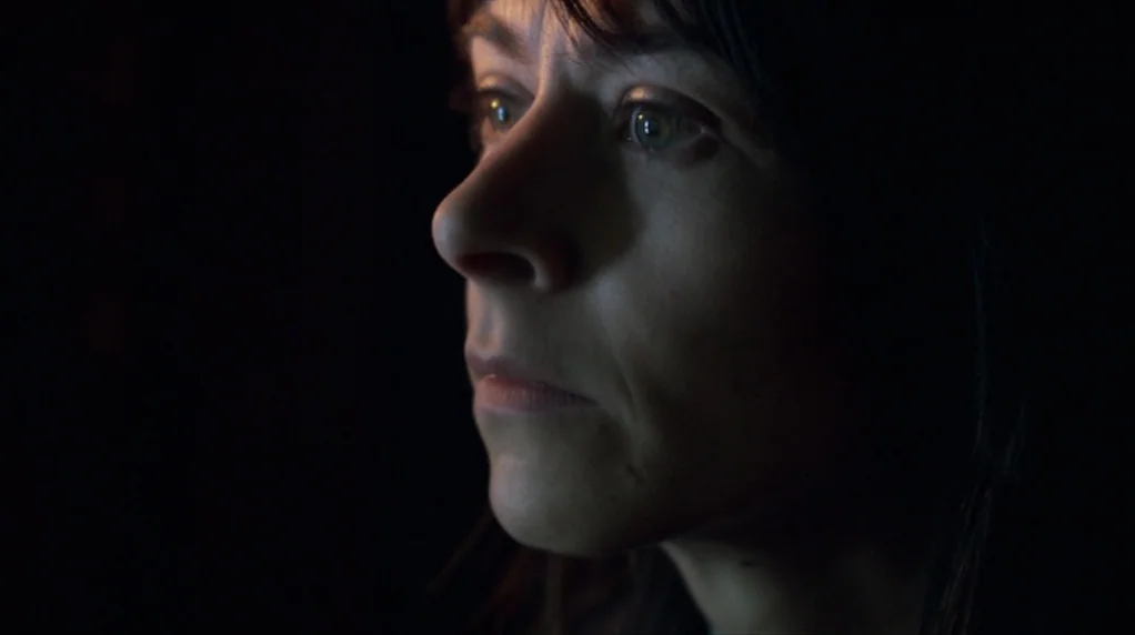 Red Road (2006) movie still: closeup — A woman's face is illuminated from the side within a pitch-black environment. She stares forward while light…