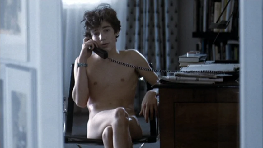 Benny’s Video (1992) movie still: medium — A home office space includes a wooden desk with a stack of books. A young man sits naked on a chair using…