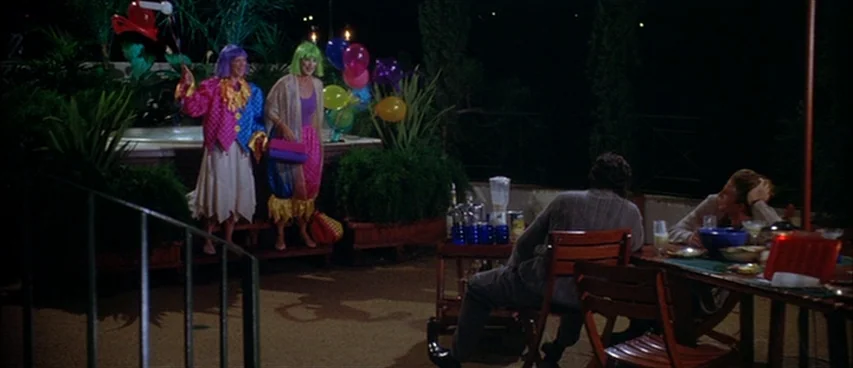 Short Cuts (1993) movie still: wide — A nighttime patio setup with potted greenery and a table holding drinks. Two people in clown-like costumes…