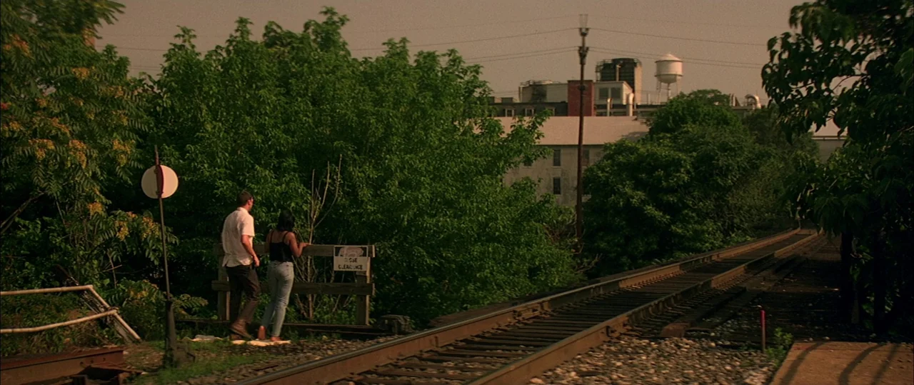 George Washington (2000) movie still: wide — A man and woman stand near a wooden barrier beside rusted railroad tracks outdoors. Dense green foliage…