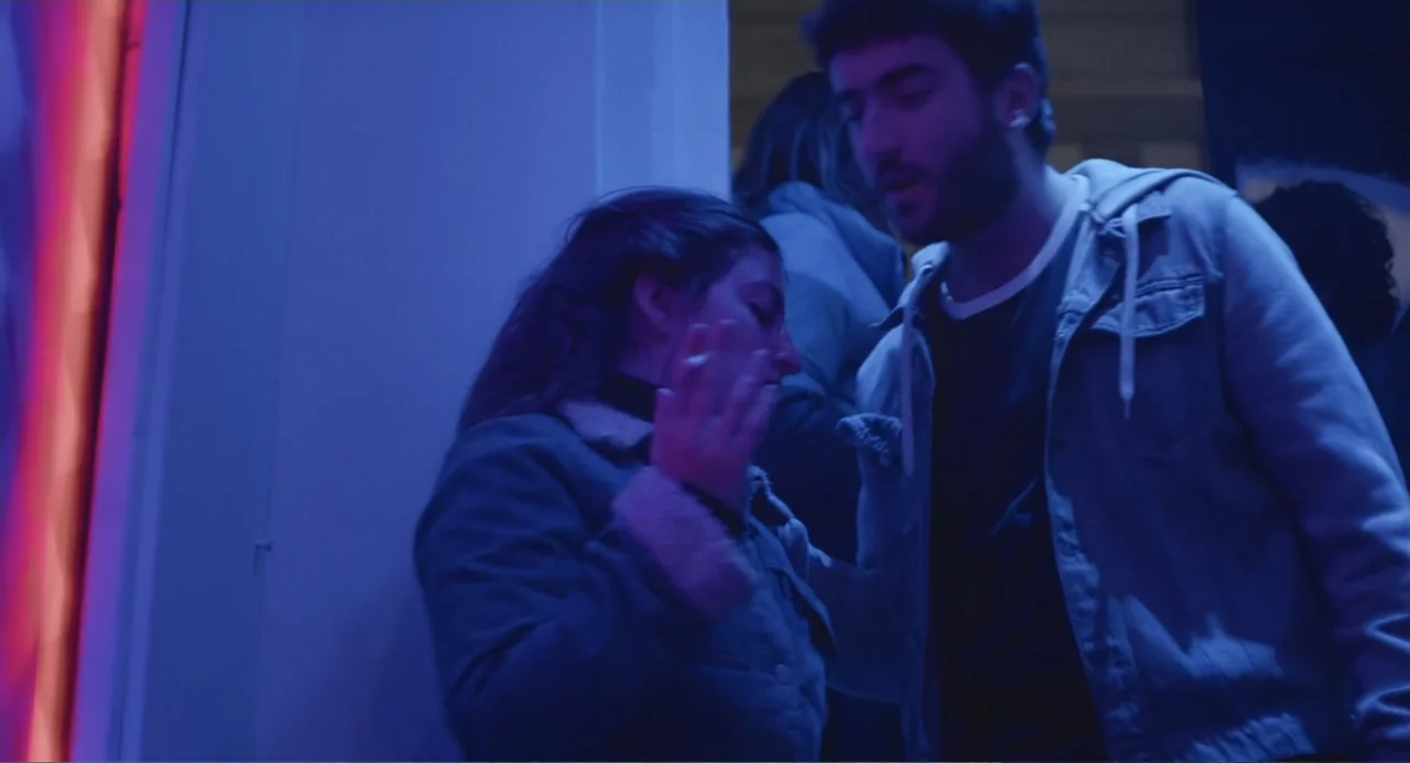 An Odd Turn (2024) movie still: medium — A dark indoor space features a red neon light strip along a doorway frame. A man and a woman stand together…