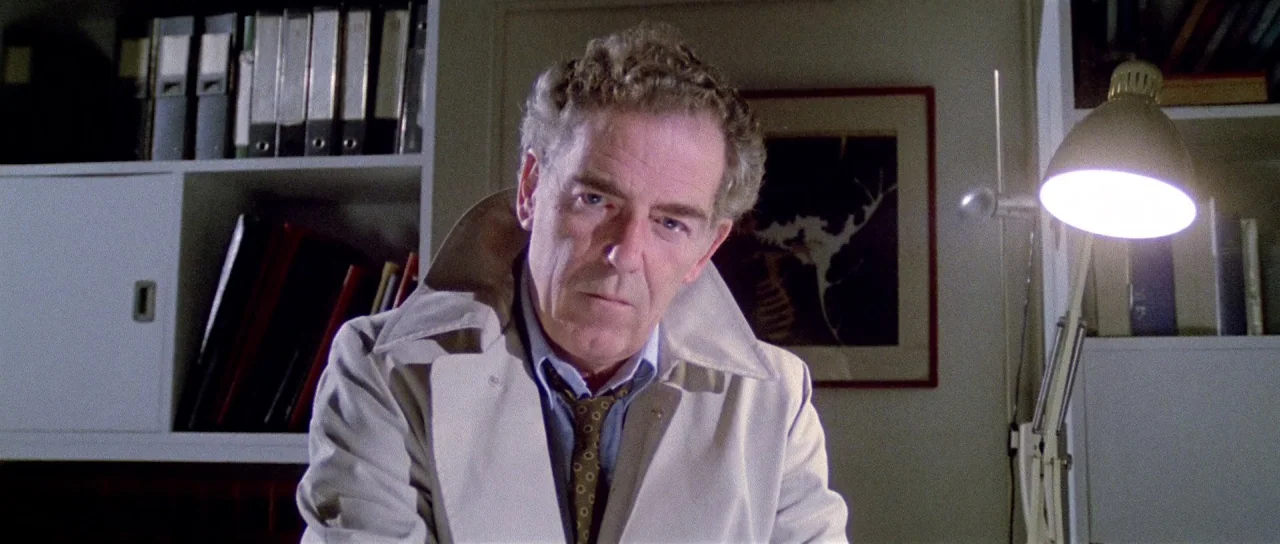 The New York Ripper (1982) movie still: medium — A study filled with shelves of ring binders and a framed print on the wall. A man sits at his desk…