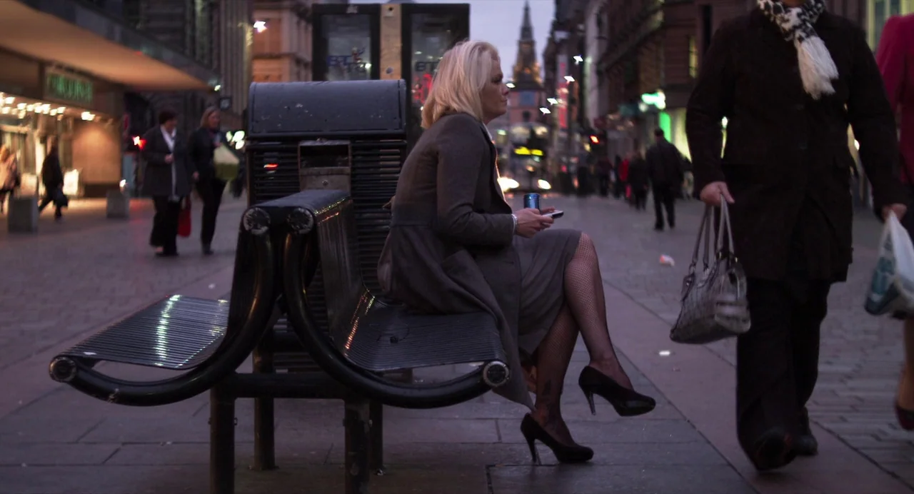 Under The Skin (2013) movie still: medium — A paved pedestrian area at twilight features a metal bench under a street light. A blonde woman sits…