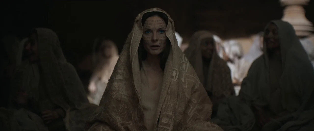 Dune: Part 2 (2024) movie still: medium — A dimly lit interior space with several figures wearing hooded, textured garments. A central woman with…