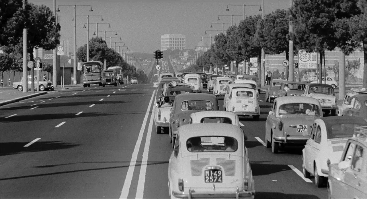 Il Boom (1963) movie still: wide — A wide multi-lane road features heavy automobile traffic moving away from the camera. The distant horizon…