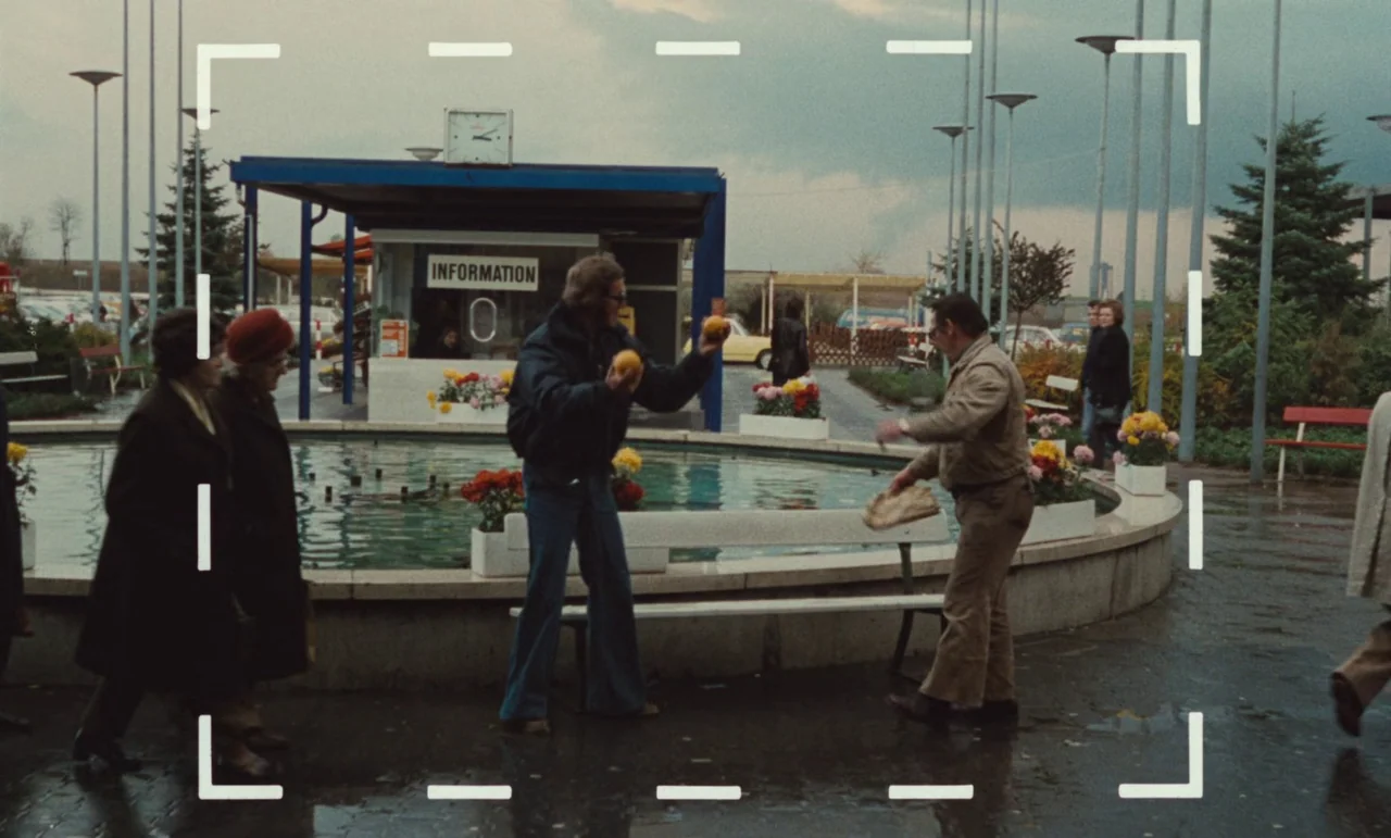 Wrong Move (1975) movie still: wide — A wet paved outdoor area features a circular stone fountain with floral planters. Two men stand on opposite…