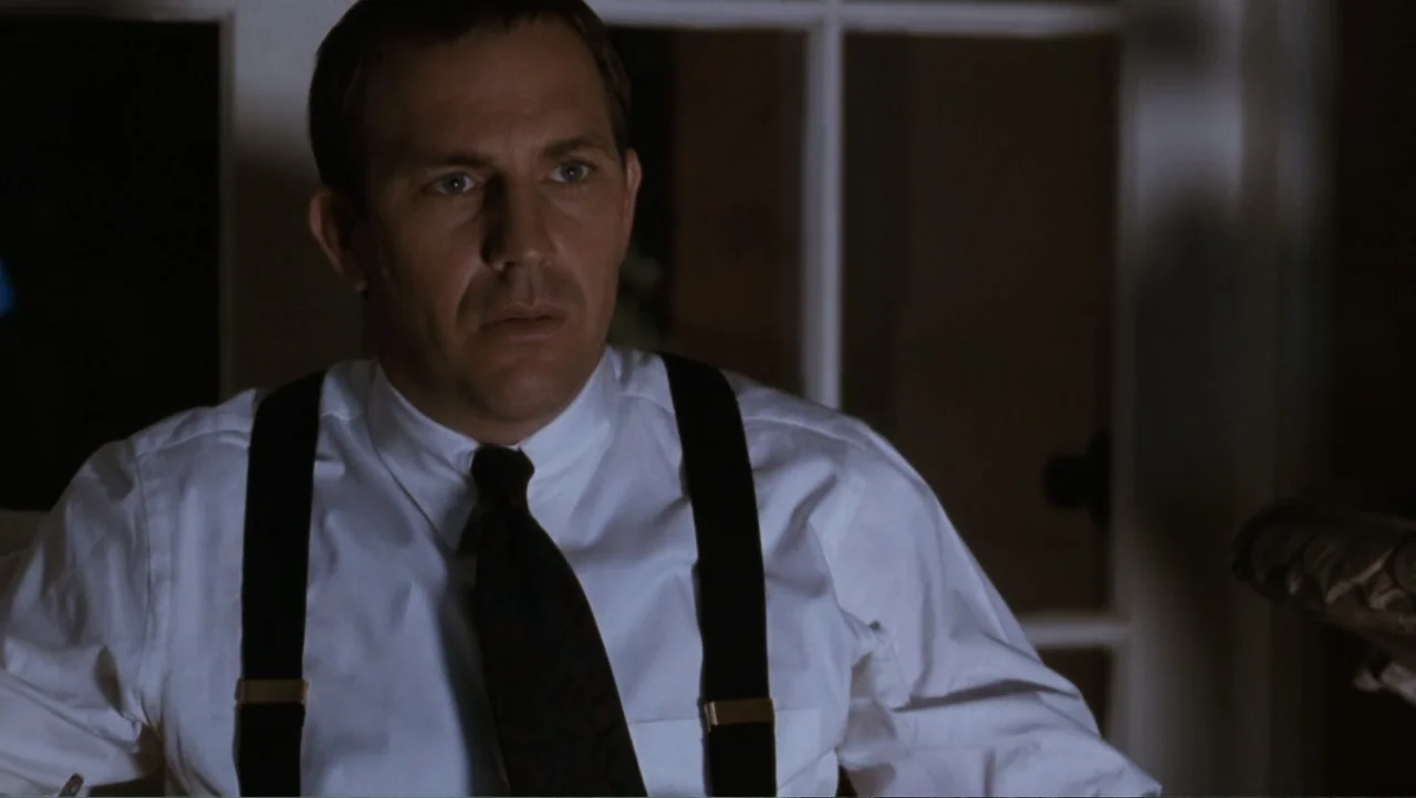 The Bodyguard (1992) movie still: medium — A dimly lit room features a window with white frames in the background. A man wearing a white shirt and…