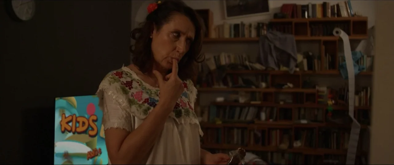 Can't Quit Mom (2022) movie still: medium — A dimly lit living room with wooden bookshelves filled with books and a television. A woman in an…