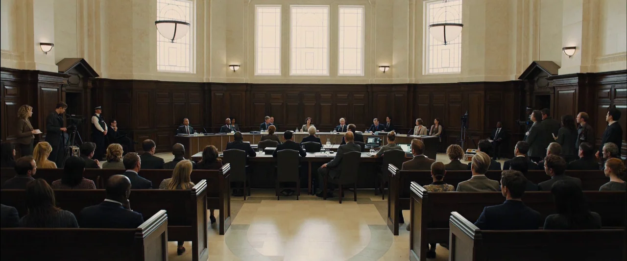 Skyfall (2012) movie still: wide — A spacious courtroom features dark wood paneled walls and high windows with natural light. People sit at a long…