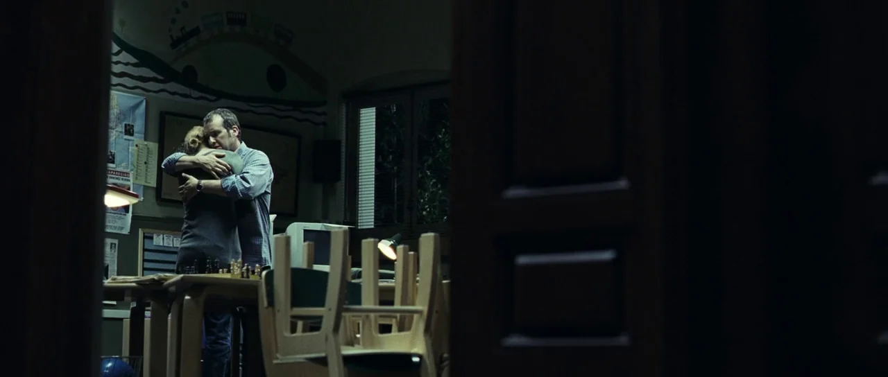 The Orphanage (2007) movie still: wide — A dark, dimly lit classroom is viewed through a partially obscured doorway. A man and a woman stand by a…