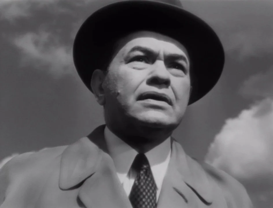 The Stranger (1946) movie still: closeup — A man stands outdoors under a sky with patchy clouds. He looks slightly upward toward something out of…