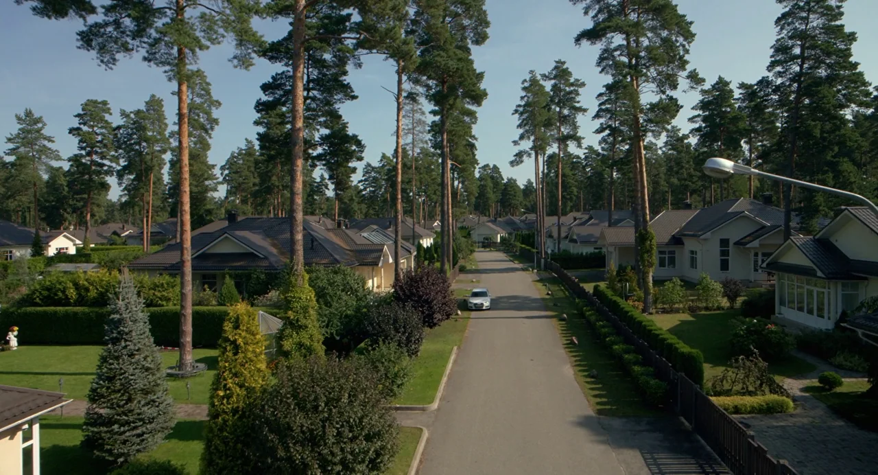 Hatching (2022) movie still: establishing — A quiet residential street is lined with manicured lawns and tall coniferous trees. A lone white sedan…
