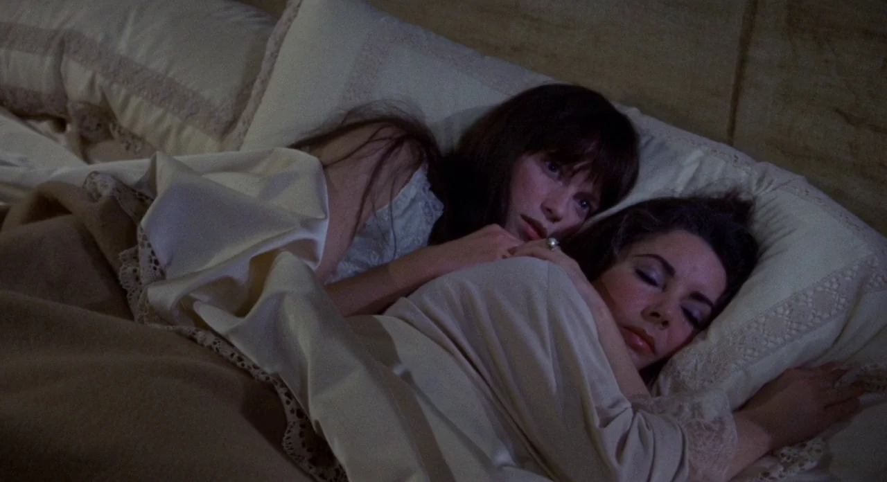 Secret Ceremony (1968) movie still: medium — Two women lie together in a bed with cream-colored bedding and lace-trimmed pillows. One woman rests…