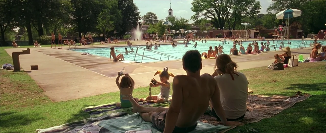Little Children (2006) movie still: wide — A bustling public swimming pool area with numerous people swimming and lounging on towels. A family of…