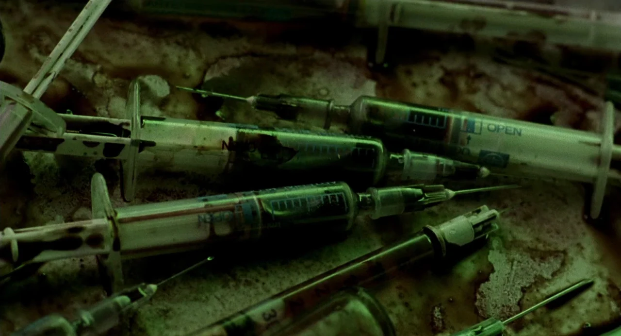 The Nameless (1999) movie still: closeup — Multiple used medical syringes are cluttered on a metal tray stained with dark residue. The syringes are…