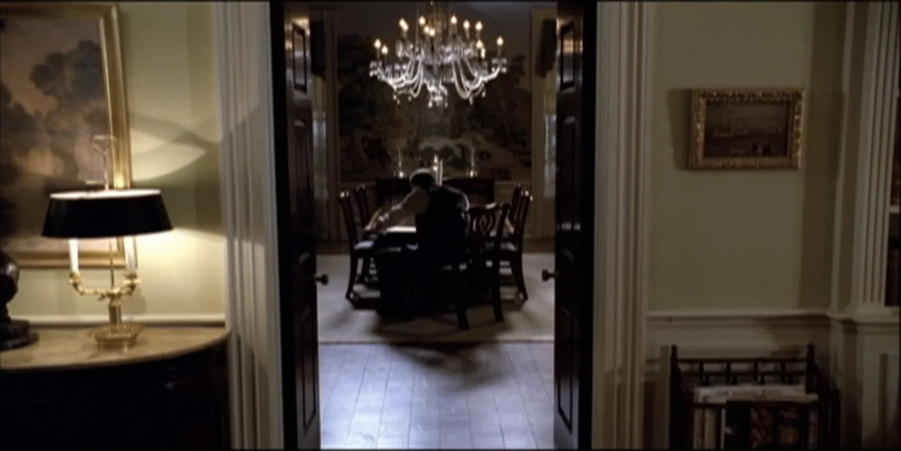 Path to War (2003) movie still: medium — A dark, formal room features a crystal chandelier hanging above a polished wooden table. A man in a dark…