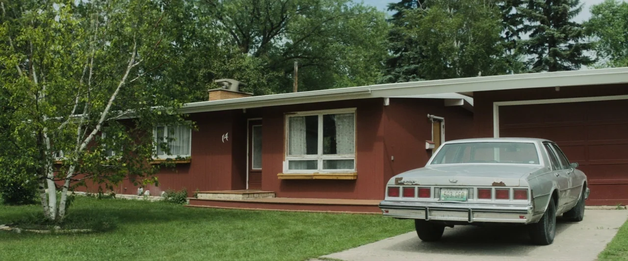The Grudge (2020) movie still: wide — A single-story red suburban house with a flat roof features a detached garage. A silver sedan is parked in the…