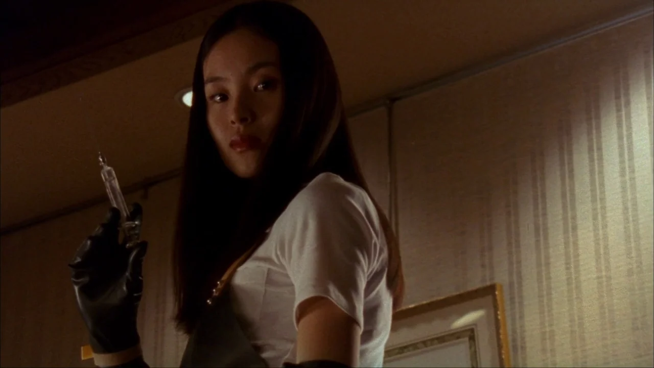 Audition (1999) movie still: medium — An interior space features patterned beige wallpaper and a framed picture on the wall. A young woman turns…