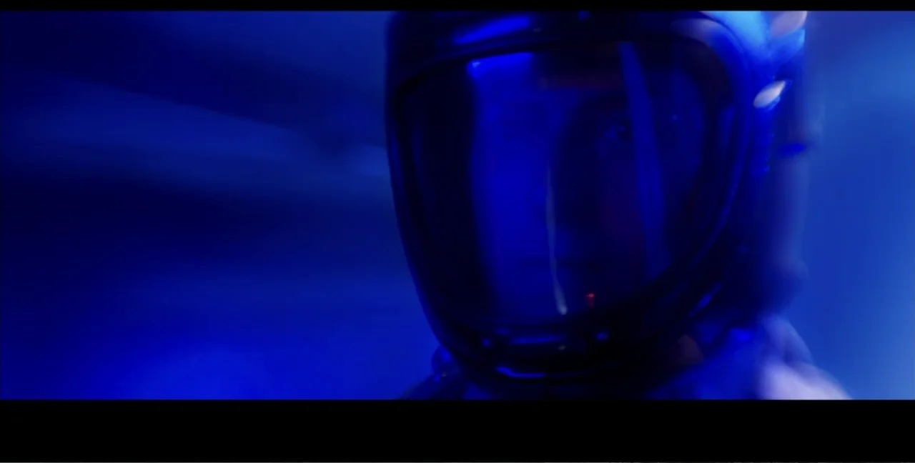 Alien Abduction: Travis Walton (2022) movie still: closeup — A person wears a helmet inside a dimly lit interior with blue light cast. The subject…