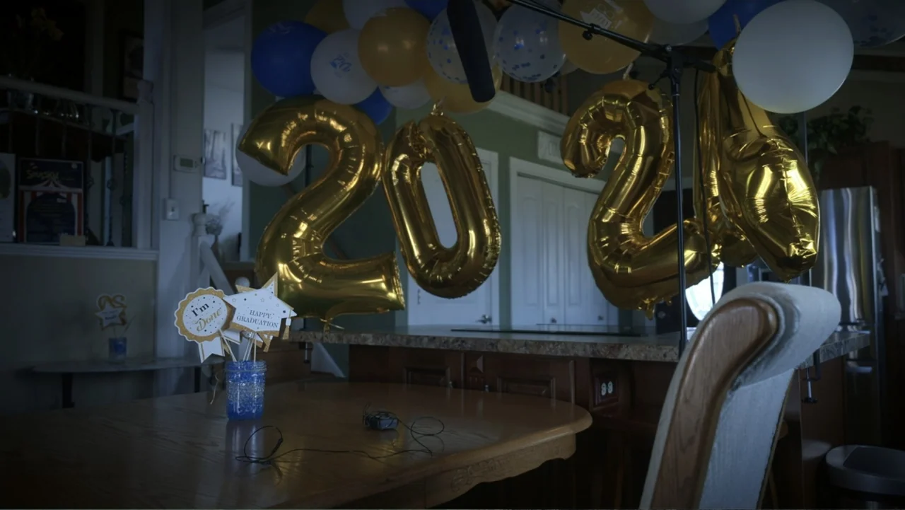Predators (2010) movie still: wide — A dining area contains a wooden table decorated with graduation signs and gold balloons. Large metallic gold…