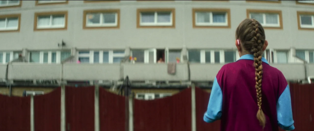 Scrapper (2023) movie still: medium — A concrete residential apartment block features rows of windows and a small balcony. A young girl stands with…