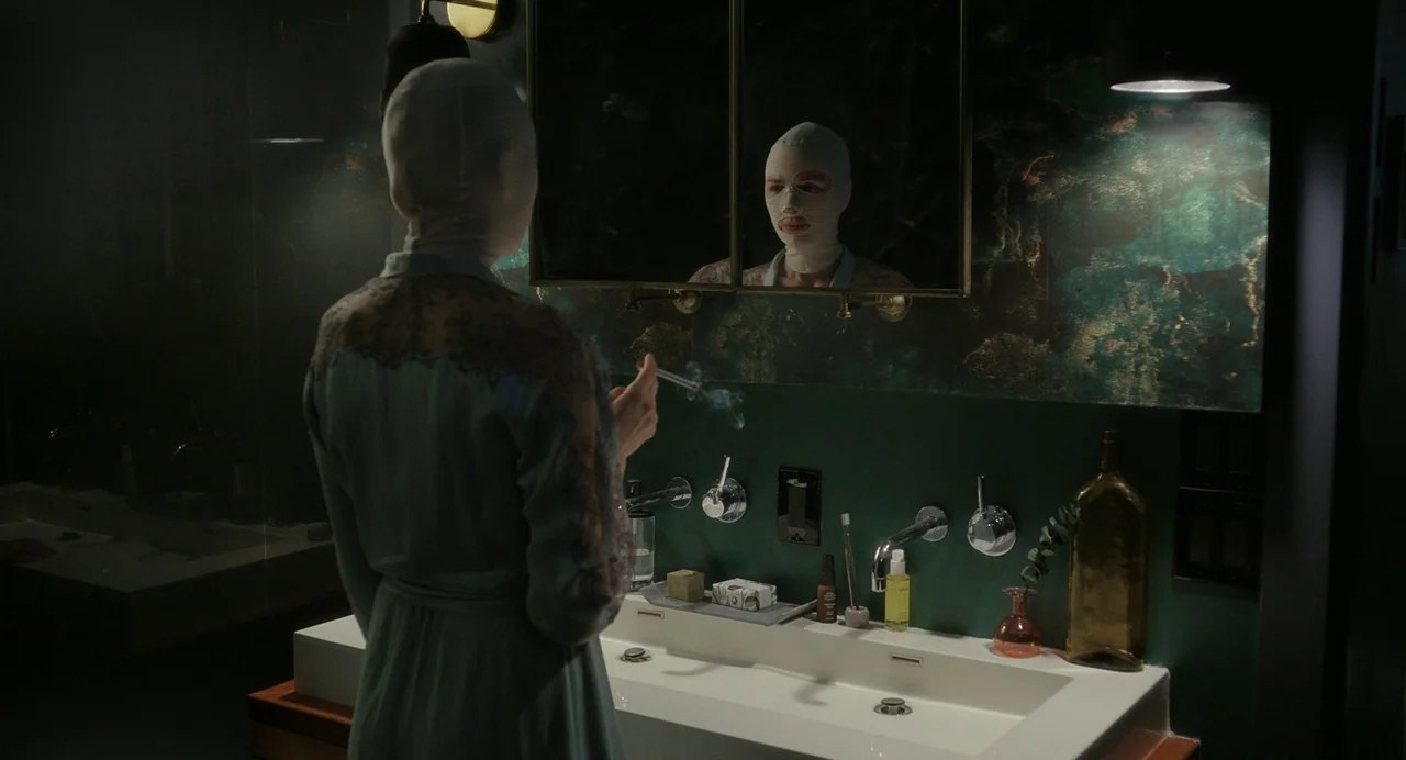 Goodnight Mommy (2022) movie still: medium — A dimly lit room with dark green walls features a modern white sink vanity. A bandaged figure stands…