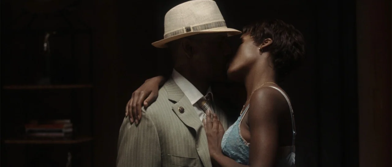 Inside Man (2006) movie still: medium — A dark indoor setting features a couple standing close in a dimly lit area. The man wears a suit and fedora…