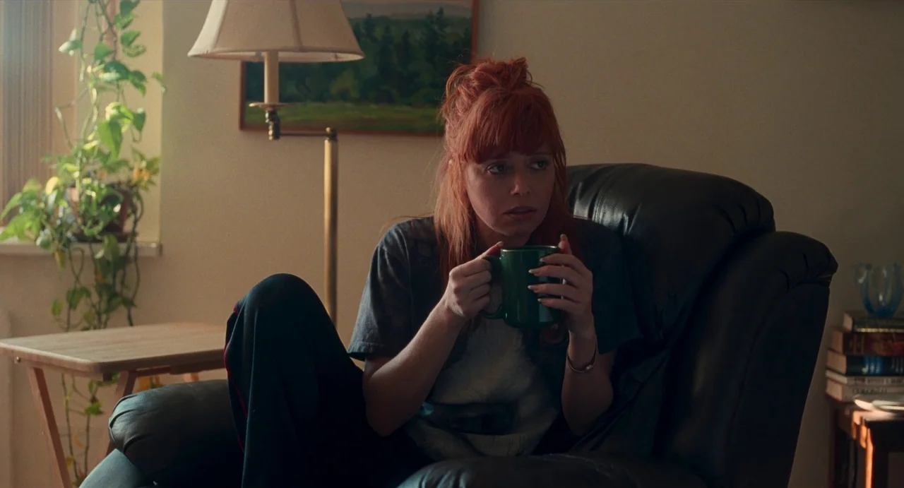 His Three Daughters (2023) movie still: medium — A woman sits in a dark leather armchair inside a residential room. She holds a green mug with both…
