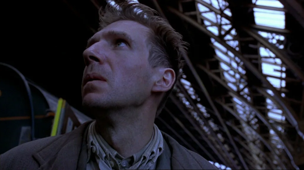 Spider (2002) movie still: closeup — A vast train station with a high glass and steel structural roof overhead. A man in a heavy coat looks upward…