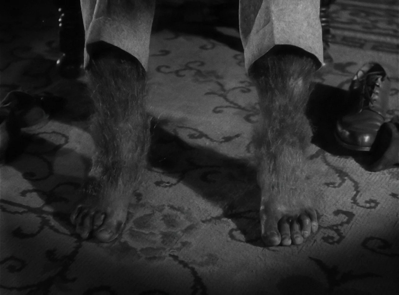 The Wolf Man (1941) movie still: closeup — A dark patterned carpet floor features a pair of hair-covered human legs and feet. The character stands…
